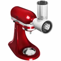 Accesorio cortador y rallador de verduras blanco acoplado al frente de una batidora KitchenAid roja sobre fondo blanco.