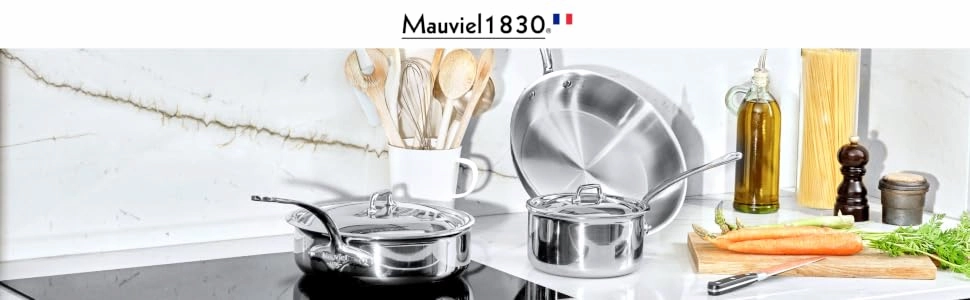 Juego de utensilios de cocina de acero inoxidable Mauviel M'Cook de 5 capas sobre una encimera de cocina con ingredientes.