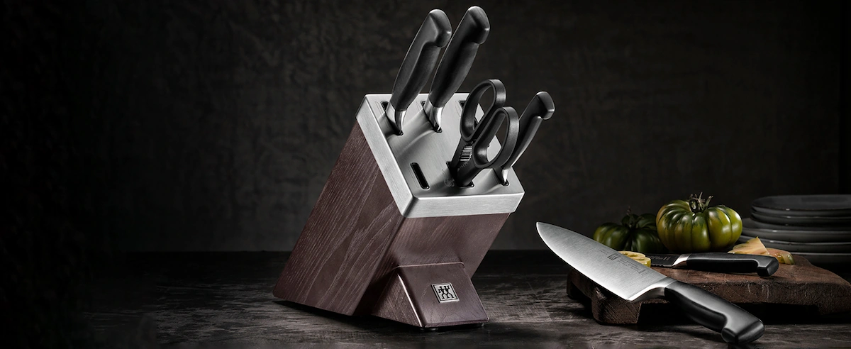 Toma panorámica con fondo oscuro y texturizado que expone el bloque de cuchillos Zwilling Four Star integrado en un ambiente culinario profesional con hortalizas frescas.