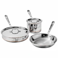 Juego de utensilios de cocina All-Clad Copper Core de 5 piezas en acero inoxidable sobre fondo blanco, incluyendo sartenes, olla y tapas con detalle de línea de cobre.